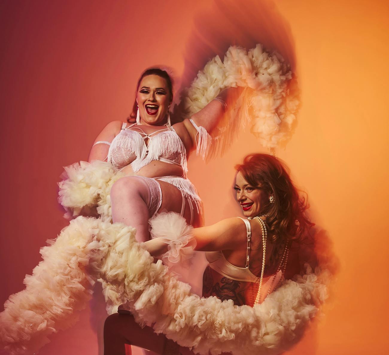 Two burlesque beauties pose together in front of an orange background. They are both dark haired and wearing white burlesque outfits with beads and fringe. They are both holding white fluffy boas that they wrap around themselves.